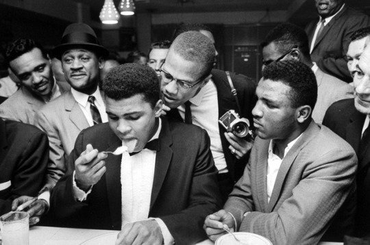 Muhammad Ali eating ice cream with Malcolm X standing over him at the Hampton House Motel 1964