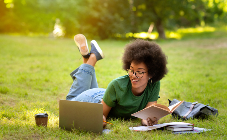 black girl laying in the grass studying on the computer and reading a book